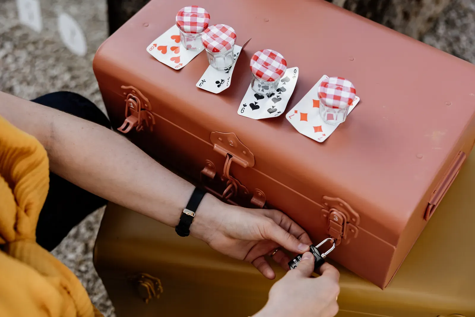 Hands working a combination padlock on a metal trunk, playing cards and jars set out on the lid