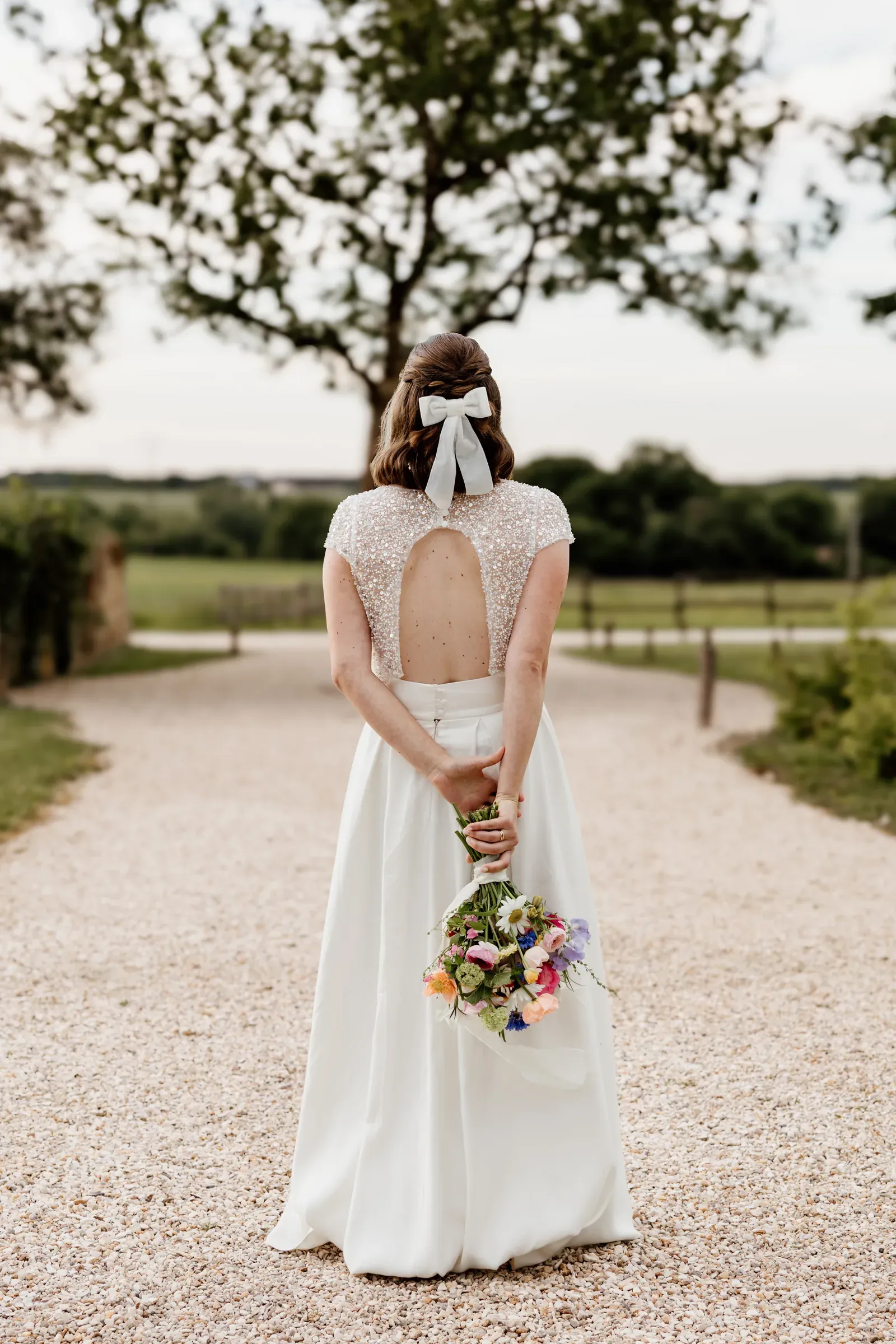 The dress code worn, seen from behind on a gravel path: a sequinned open-back top, a long white skirt, a white hair bow and a wildflower bouquet held low