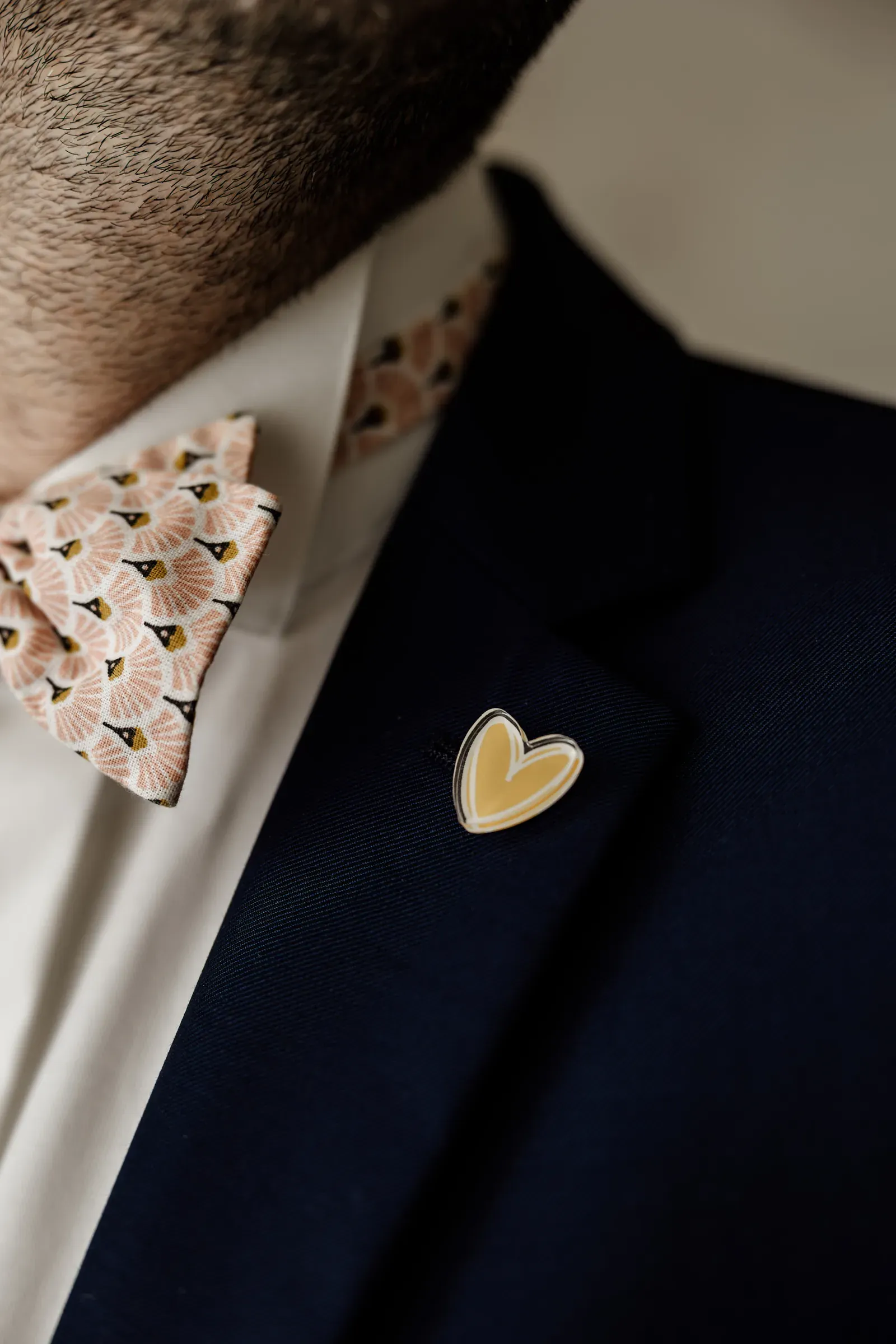 Enamel heart pin in the identity's ochre, worn on a navy lapel beside a bow tie in the scalloped pattern