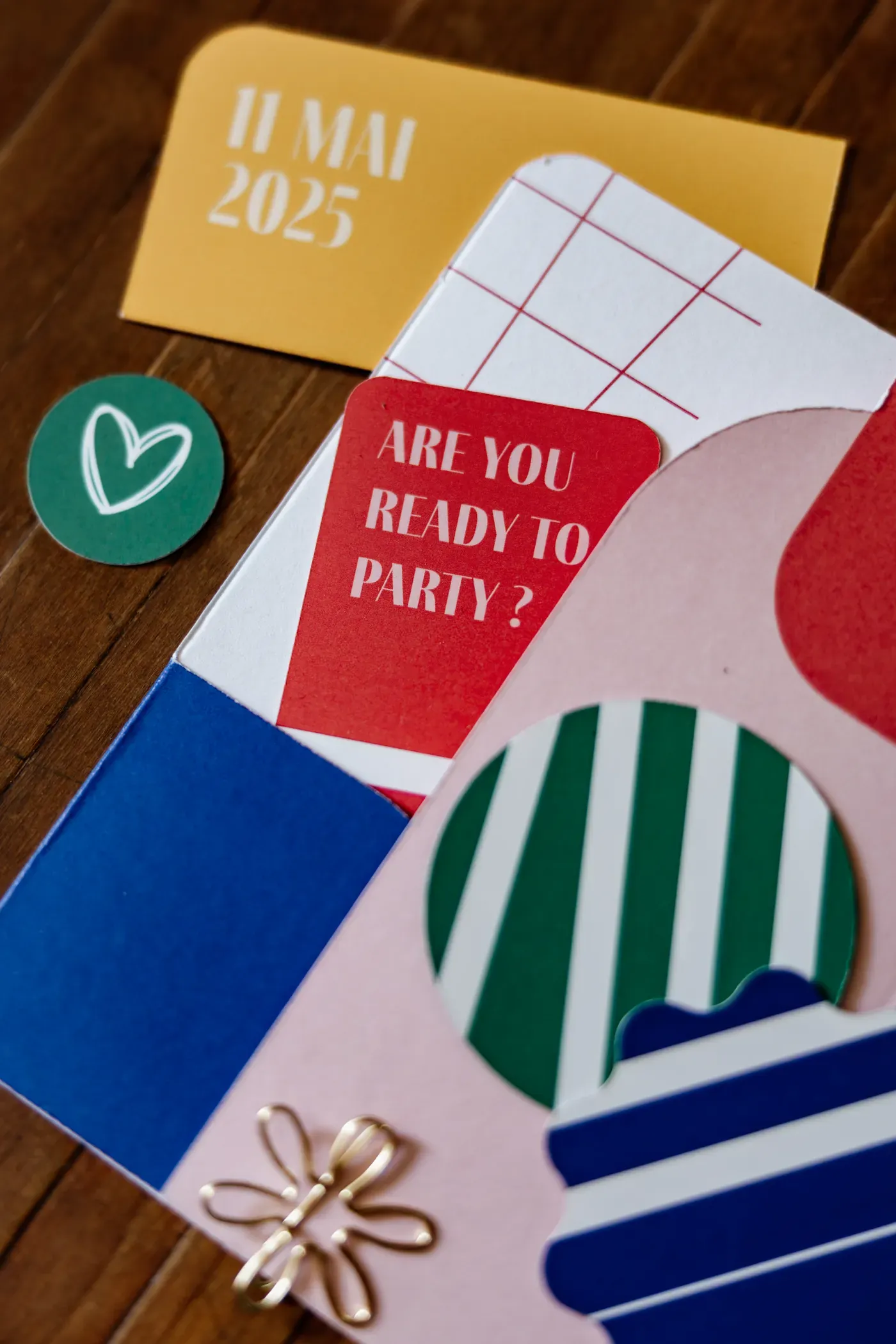 Close view of the printed invitation: die-cut cards, coloured blocks and a heart sticker on a wooden table