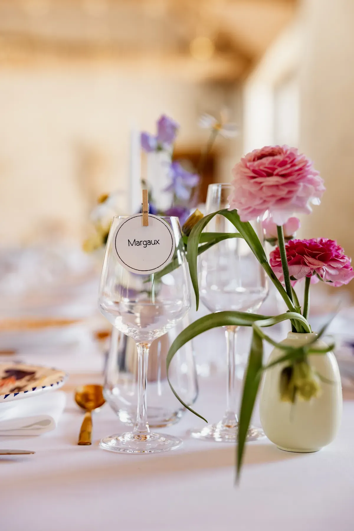 Place card clipped to a wine glass on the laid table, beside a small vase of ranunculus