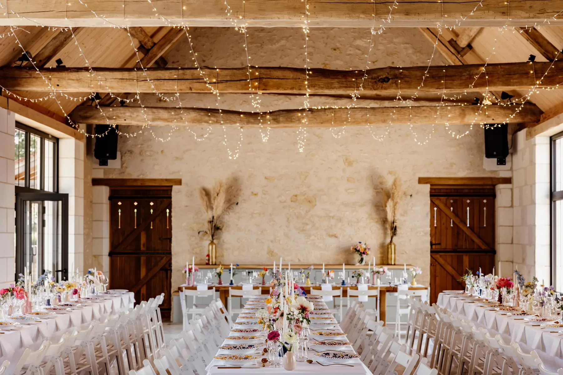 Reception hall under exposed beams and string lights, long tables laid for a hundred guests
